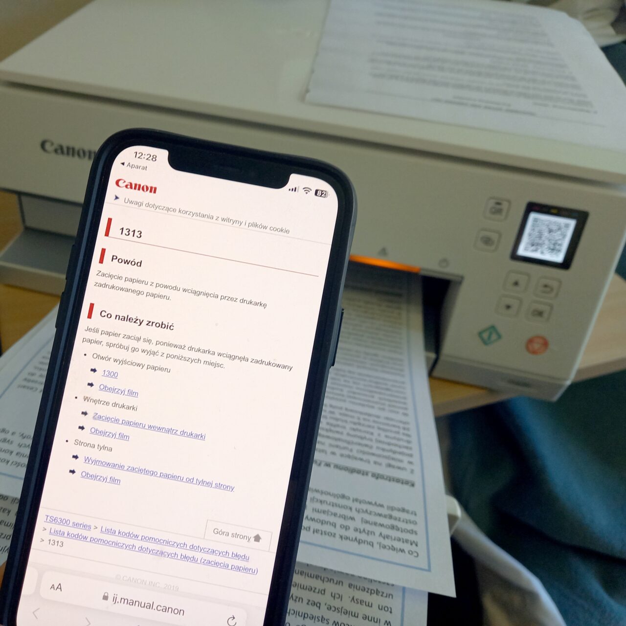 Smartfon wyświetlający stronę pomocy technicznej z instrukcjami dotyczącymi naprawy drukarki Canon, widocznej w tle na biurku z papierami.