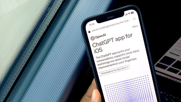 Smartfon trzymany w ręce z wyświetloną stroną OpenAI promującą aplikację ChatGPT dla iOS, w tle rozmyte elementy komputerowe.