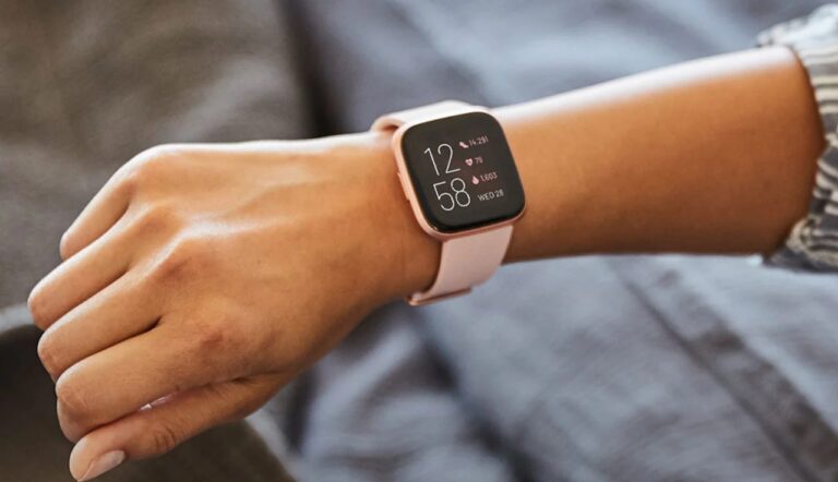 Ręka osoby z założonym na nadgarstku inteligentnym zegarkiem o różowym pasku, wyświetlającym czas i dane zdrowotne.