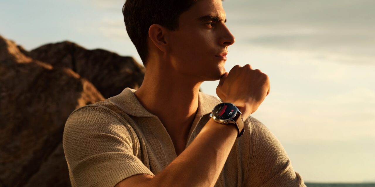 Młody mężczyzna patrzy w dal, nosząc inteligentny zegarek na nadgarstku; tło z skalistym terenem i niebem w czasie zachodu słońca.