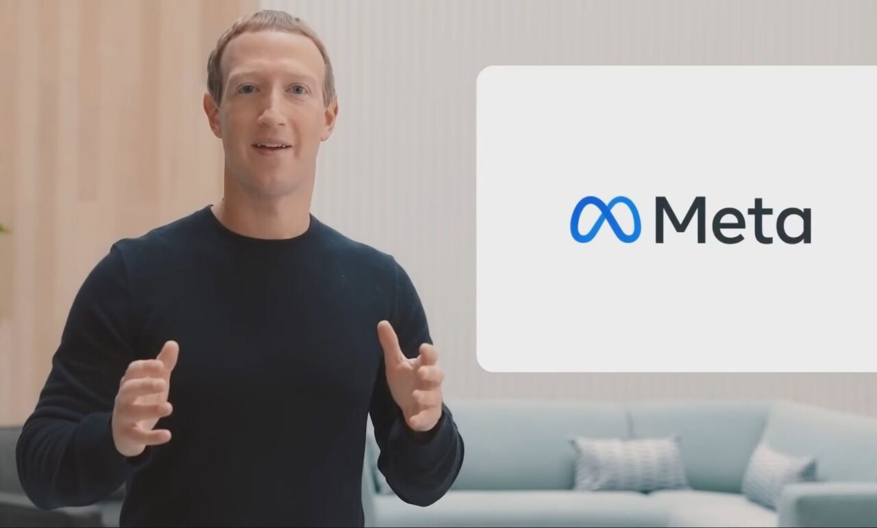 Mężczyzna w czarnej koszulce gestykuluje, w tle logo firmy Meta na ekranie obok niebieskiej kanapy.