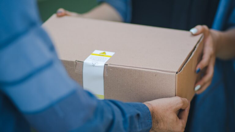Dopłata do przesyłki jako forma oszustwa. Dwie osoby przekazują sobie kartonowe pudełko, na którym widnieje etykieta ostrzegawcza.