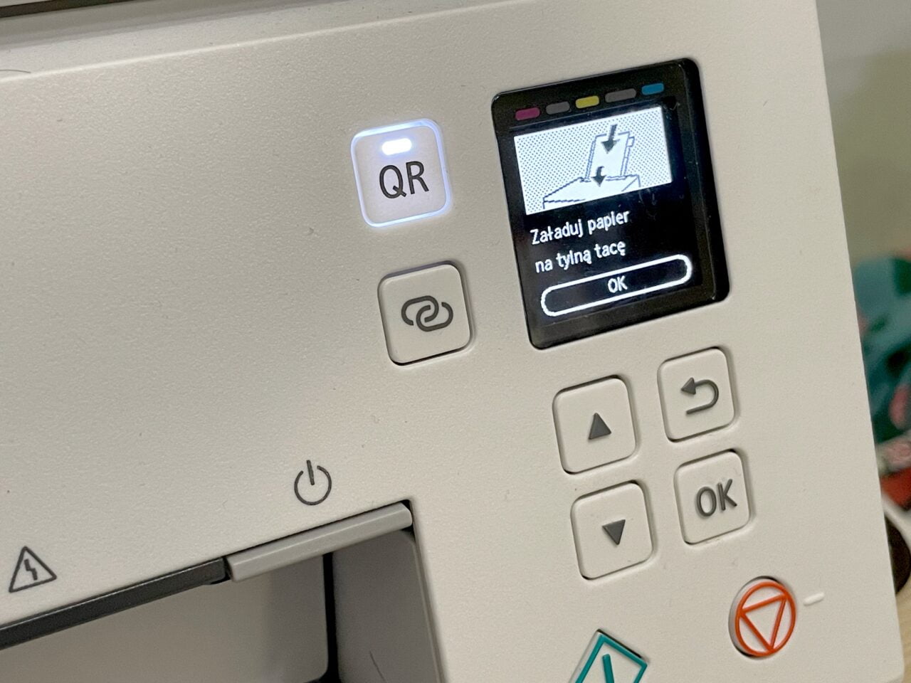 Panel kontrolny urządzenia z ekranem wyświetlającym monochromatyczny obrazek i napis "Załaduj papier na tylną tacę" z przyciskiem "OK" oraz innymi przyciskami sterującymi i symbolem QR.