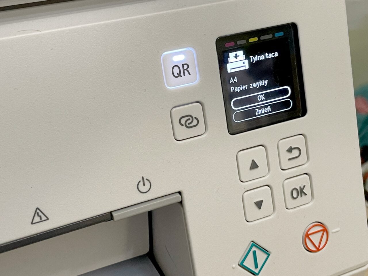 Panel sterowania drukarki z wyświetlaczem pokazującym komunikat "Tłyna taca, A4, Papier zwykły", przyciskami nawigacyjnymi oraz symbolami QR i zasilania.