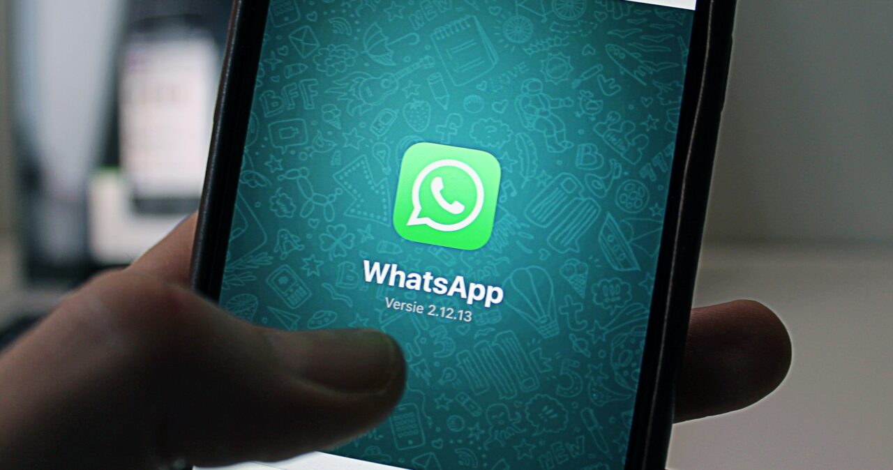 Ekran smartfona trzymanego w dłoni z otwartą aplikacją WhatsApp, wersja 2.12.13, na tle z ikonami komunikacji.