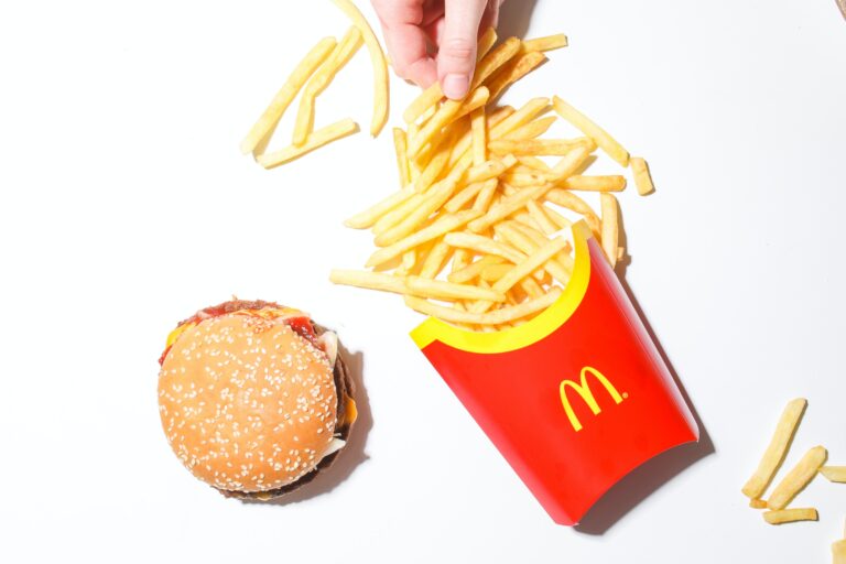 Hamburger z bułką sezamową obok czerwonego opakowania frytek z logiem McDonald's na białym tle, ręka łącząca frytki.