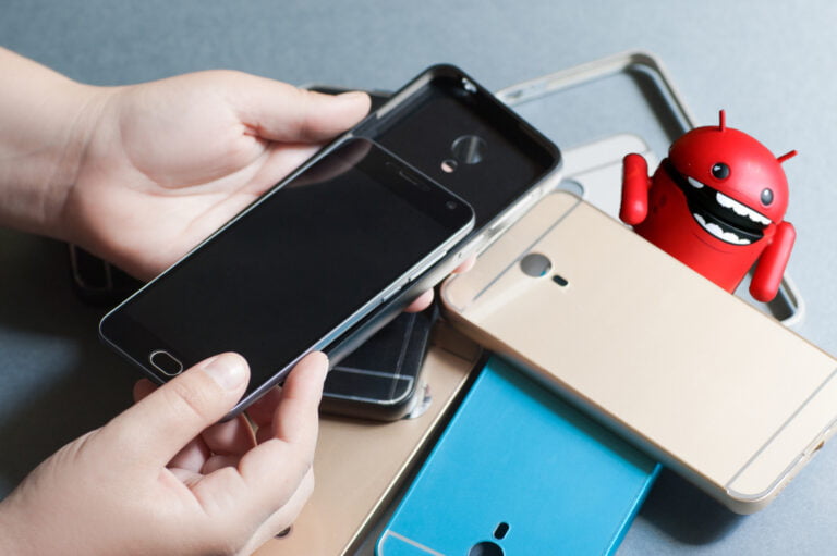 Dłonie osoby trzymające smartphone nad stosem innych telefonów komórkowych różnych marek i kolorów z zabawkowym robotem o wyglądzie Androida w tle.