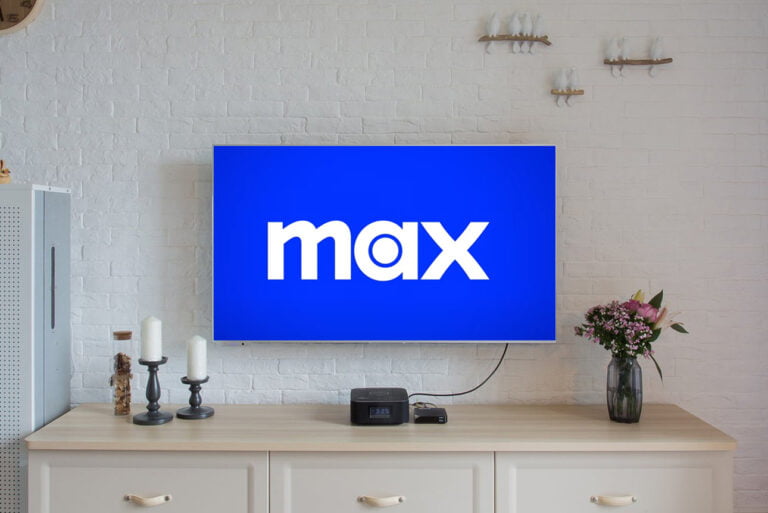 Telewizor na ściennej półce wyświetlający logo "max" na niebieskim tle, przed białą ceglaną ścianą, z bukietem kwiatów i świecami na drewnianej szafce poniżej.