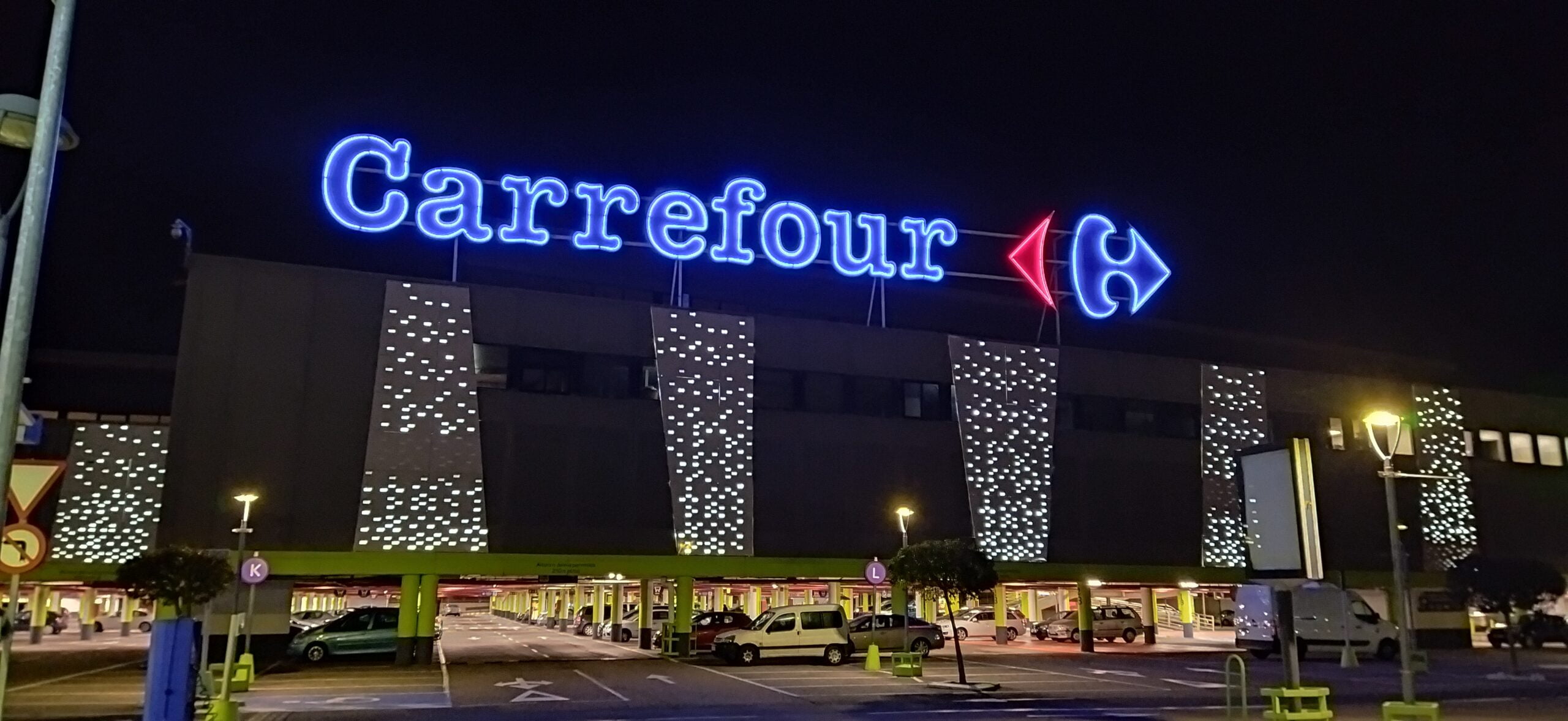 Parking przed budynkiem supermarketu Carrefour wieczorem, z naświetlonymi literami i strzałką logo na elewacji.