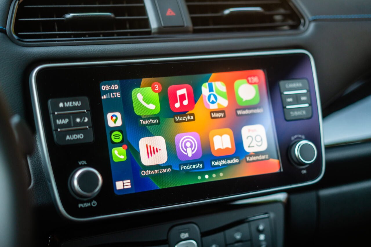 Ekran systemu multimedialnego w samochodzie wyświetlający kolorowe ikony aplikacji, takie jak telefon, muzyka, mapy, wiadomości, pod którymi znajdują się przyciski sterowania.