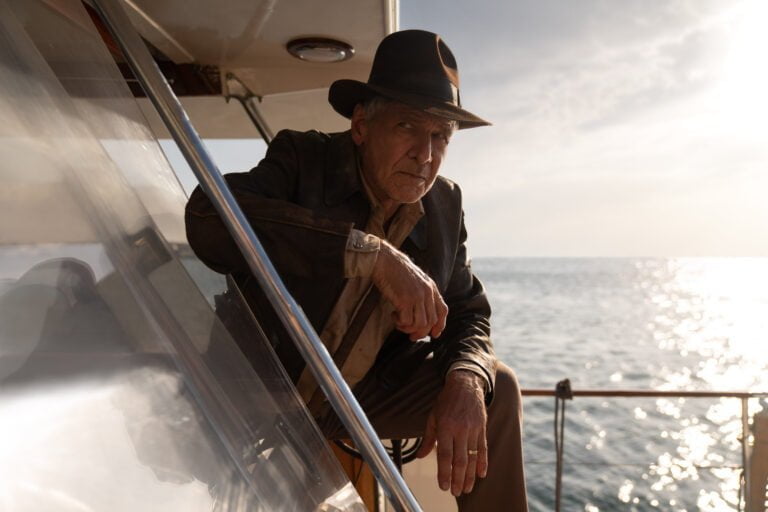 Starszy mężczyzna w czarnym kapeluszu oparty o barierkę jachtu, z tłem morza i przebłyskującego słońca.