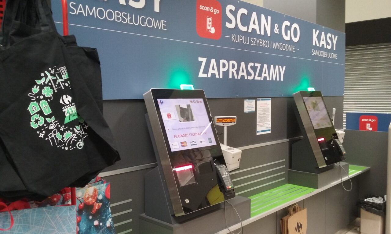 Stacja samoobsługowa w sklepie z ekranem dotykowym i terminalem płatniczym, torba na zakupy i zapakowane przedmioty obok, nad kioskiem napis "SCAN & GO KASY SAMOOBSŁUGOWE ZAPRASZAMY".