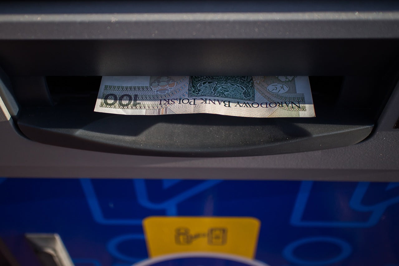 Banknot 100 złotych wystaje z wlotu bankomatu, prowadzonego przez banki.