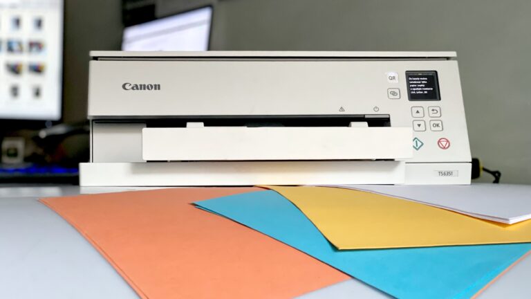 Biała drukarka atramentowa Canon na biurku z kolorowymi kartkami ułożonymi na pierwszym planie.