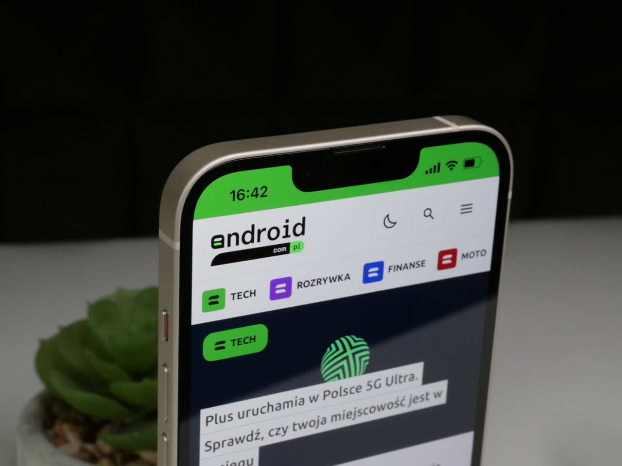 Smartfon wyświetlający stronę internetową z logo "android" i polskimi kategoriami: TECH, ROZRYWKA, FINANSE, MOTO, na tle ciemnym z rośliną w niewyraźnym tle.