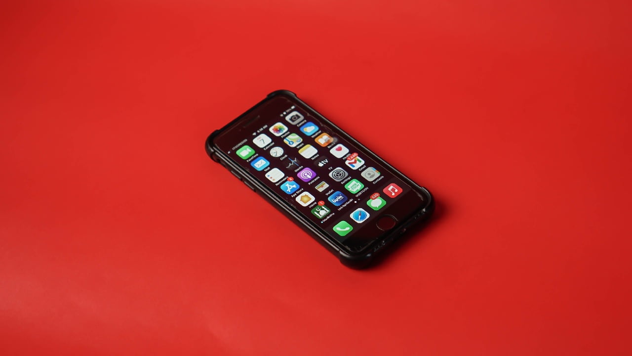 Smartfon z czarną obudową i włączonym ekranem wyświetlającym kolorowe ikony aplikacji, położony na jednolitej czerwonej powierzchni.