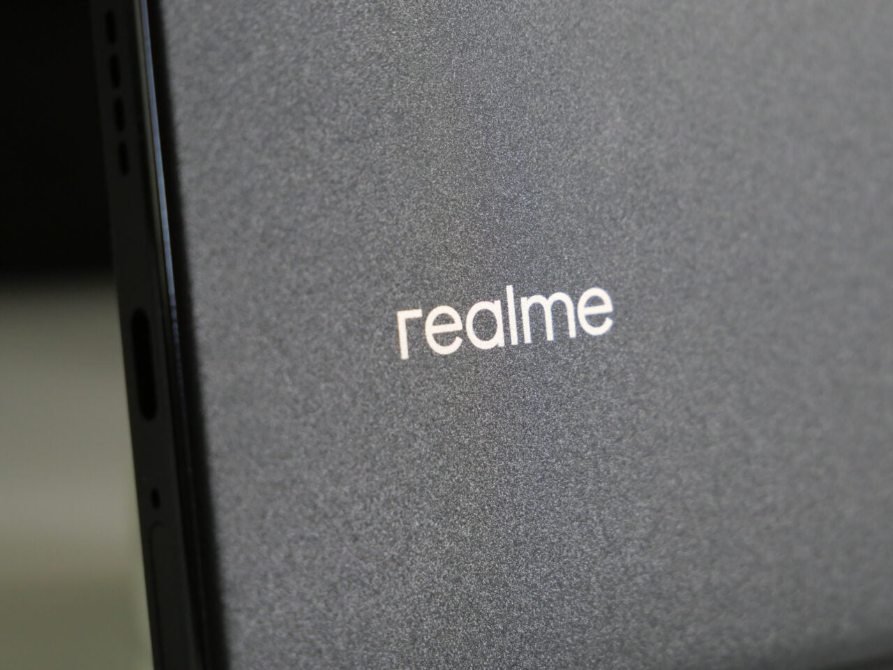 Metaliczne logo "realme" na szarej teksturowanej powierzchni.