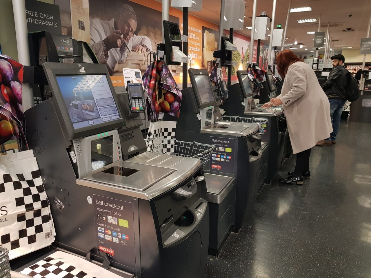 Caixas de autoatendimento em um supermercado com usuários escaneando e embalando compras.