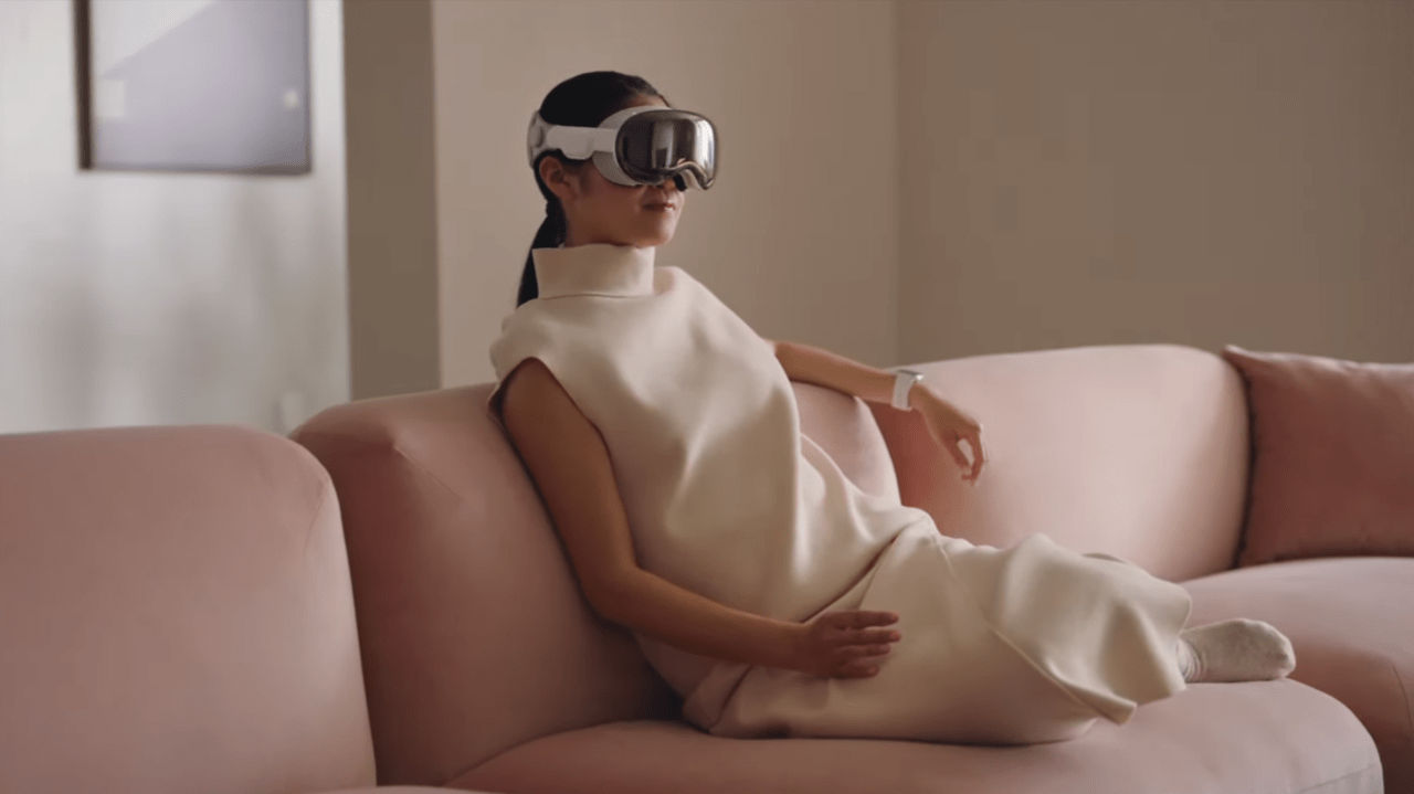 Kobieta w białej sukience siedzi na różowej sofie, ma na głowie gogle Apple Vision Pro wirtualnej rzeczywistości.