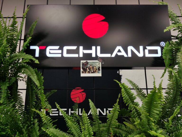 Tablica z logo "Techland" otoczona zielonymi paprociami.