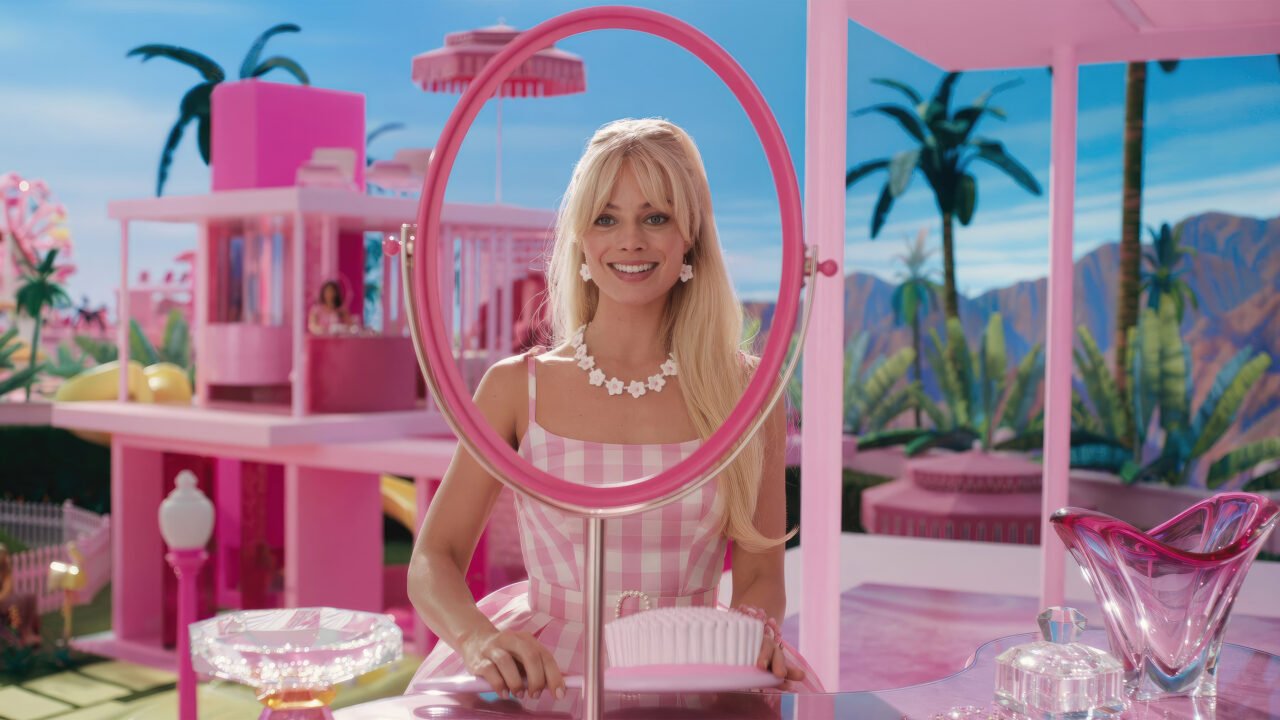 Film The Sims nabiera kształtów. Uśmiechnięta Barbie z blond włosami ubrana w różowo-białą kraciastą sukienkę, z białymi kwiatowymi akcesoriami, stoi przy różowym stoliku w intensywnie różowym pomieszczeniu z uroczymi ozdobami i widokiem na palmy oraz góry w tle.