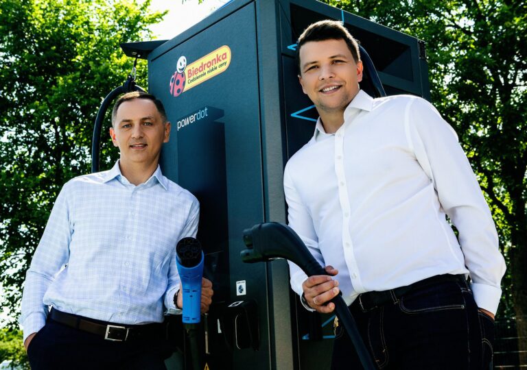 Dwóch uśmiechniętych mężczyzn w koszulach stoi przy ładowarce do samochodów elektrycznych z logo "Biedronka" i "powerdot".