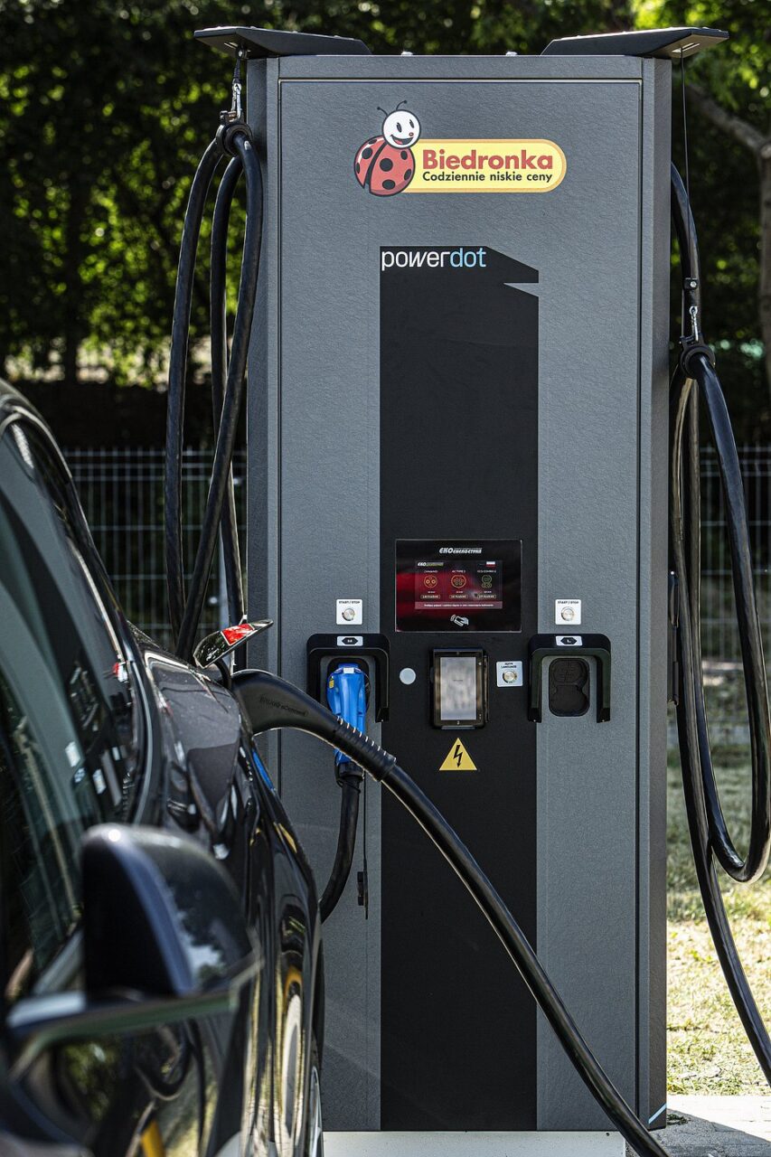 Stacja ładowania pojazdów elektrycznych marki PowerDot, z logo Biedronka, do której podłączony jest czarny samochód elektryczny.