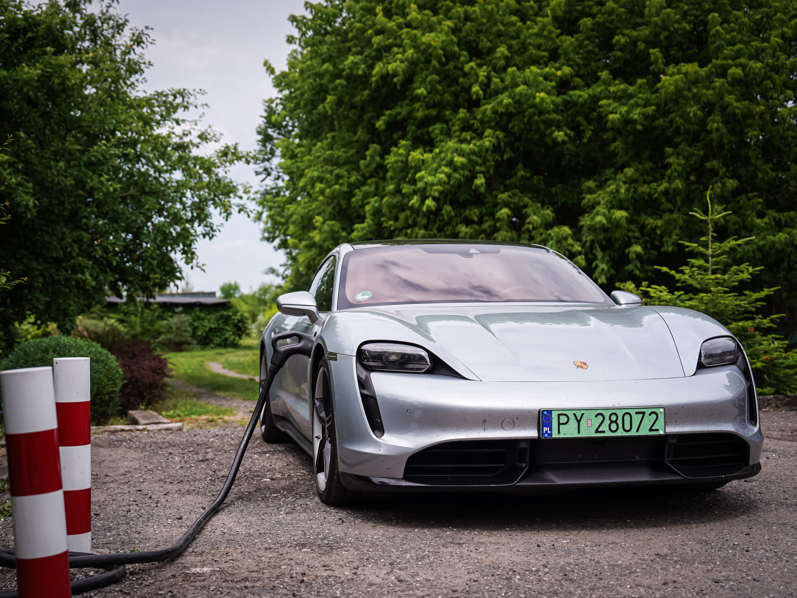 Srebrny sportowy samochód elektryczny marki Porsche podłączone do stacji ładowania na zewnątrz, otoczone zielenią.