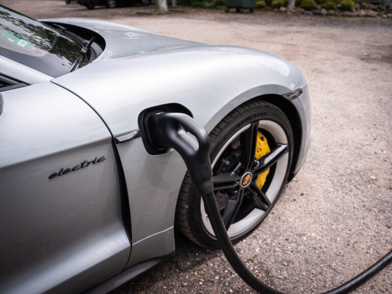 Szary samochód elektryczny ładowany kablem, z napisem "electric" na bocznej stronie pojazdu i widocznym żółtym hamulcem w tle.