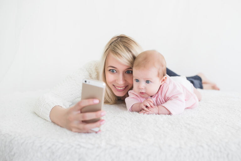 Mózg dziecka a smartfony. Uśmiechnięta kobieta w białym swetrze i małe niemowlę w różowym ubranku leżą na białym kocu i robią sobie selfie za pomocą smartfona.
