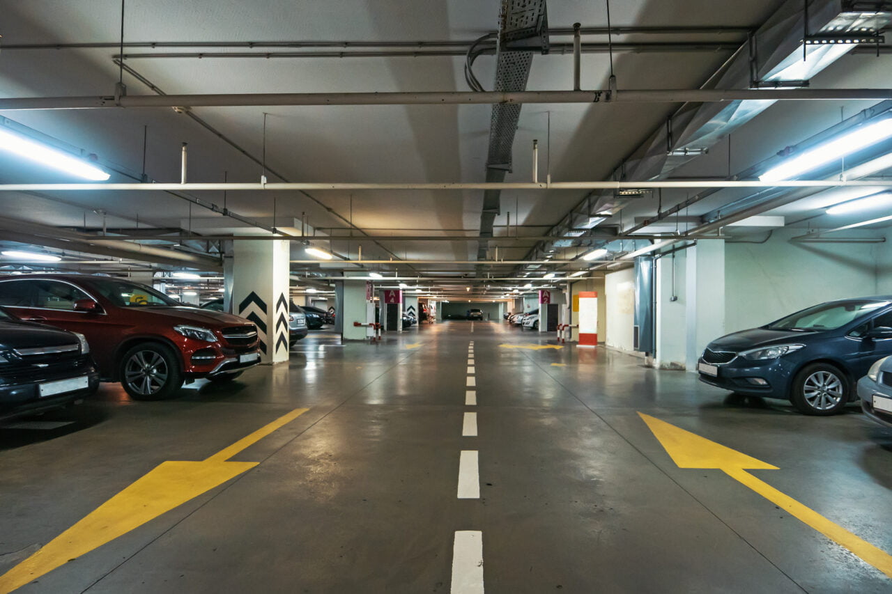 Wnętrze parkingu podziemnego z zaparkowanymi samochodami po obu stronach, oznakowanymi pasami ruchu i strzałkami kierunkowymi na podłodze. W tle może znajdować się ładowarka dla elektryków,