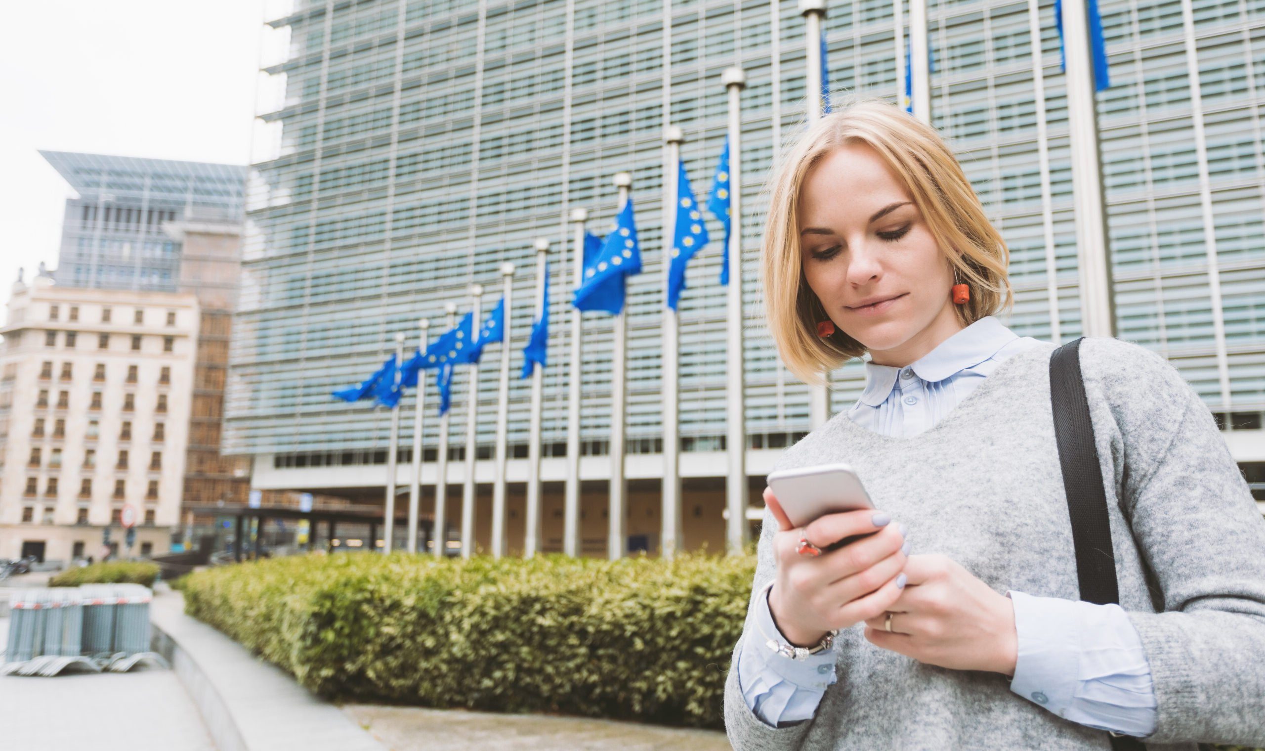 Młoda kobieta korzystająca ze smartfona na tle budynku z flagami Unii Europejskiej.