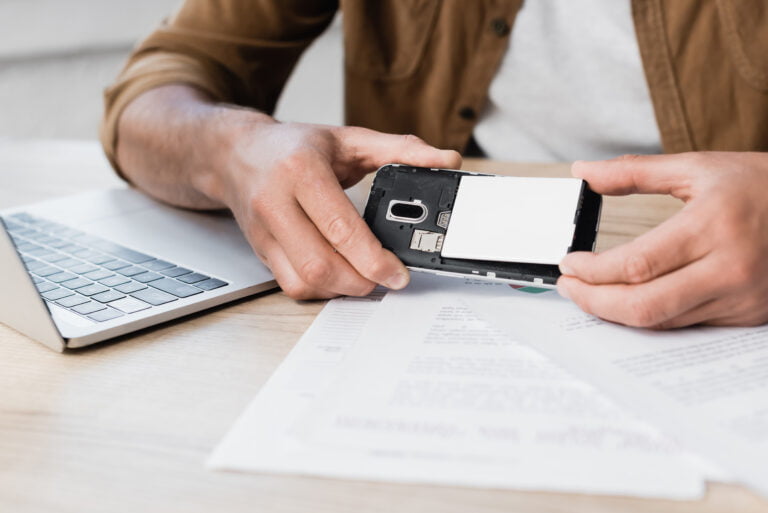 Osoba trzymająca rozebrany smartfon nad biurkiem z laptopem i dokumentami.