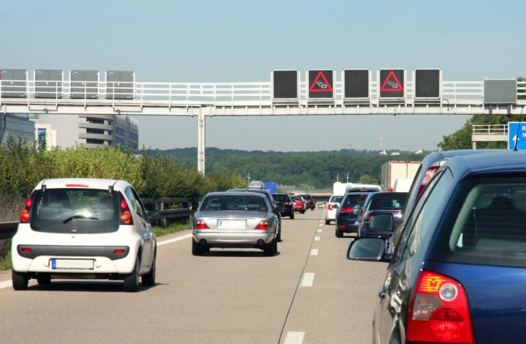 Korek na autostradzie z samochodami w kierunku trzech zamkniętych pasów ruchu, nad którymi znajdują się tablice z sygnałami ostrzegawczymi o zjeździe z drogi.