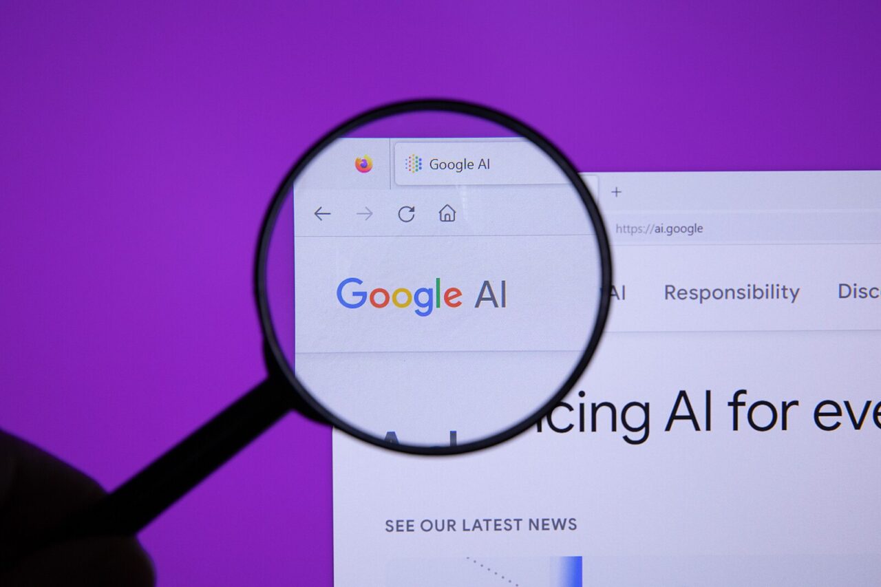 Lupa trzymana w dłoni powiększająca napis "Google AI" na ekranie monitora, na fioletowym tle.