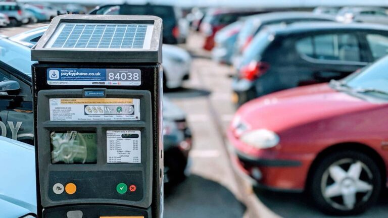 Automat do płatności za parking zasilany energią słoneczną na tle rozmytych, zaparkowanych samochodów.