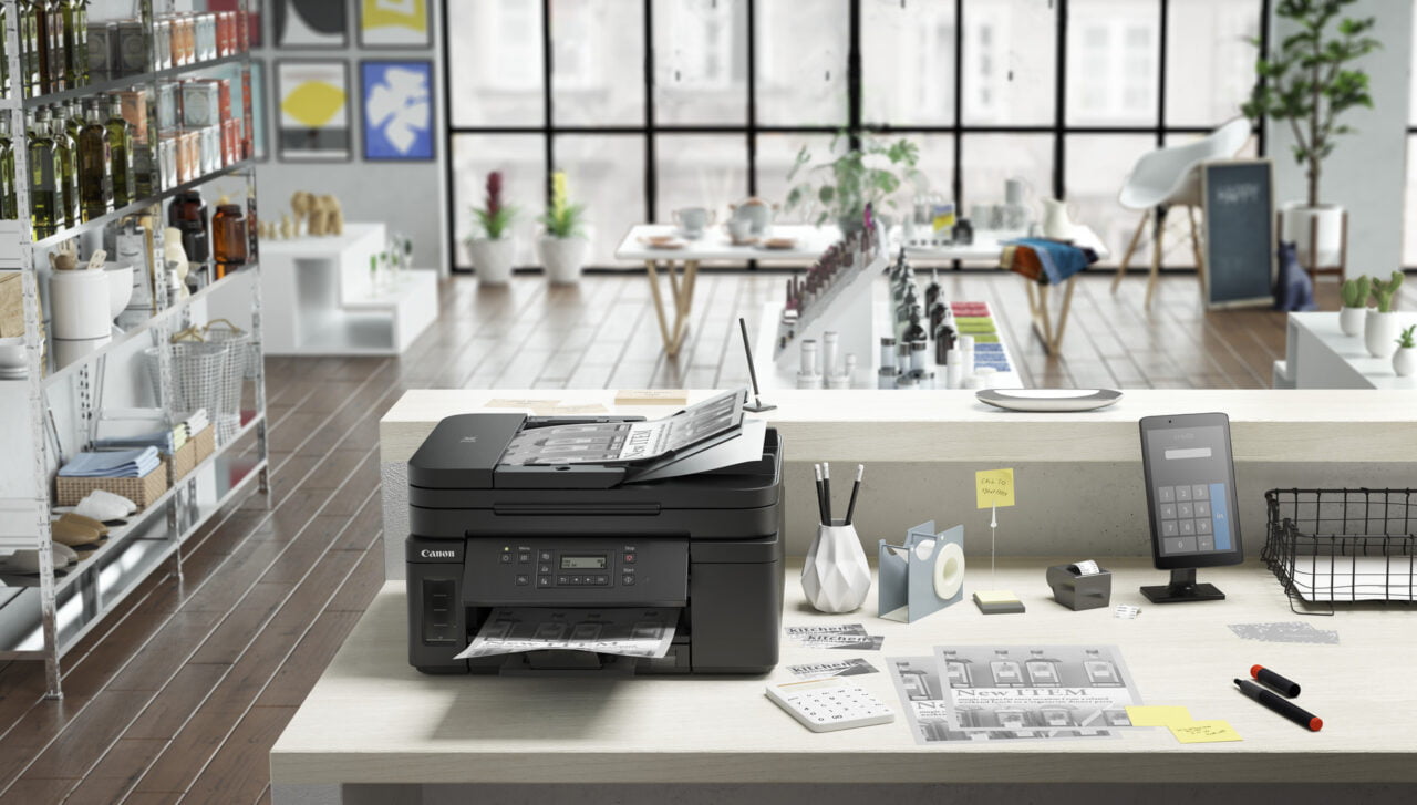 Wielofunkcyjne urządzenie biurowe marki Canon na biurku w jasnym, nowoczesnym biurze. Przy urządzeniu znajdują się drukowane materiały, długopisy i notatki samoprzylepne. W tle widoczne są regały z przyborami i rośliny doniczkowe. Drukarka Pixma - ranking