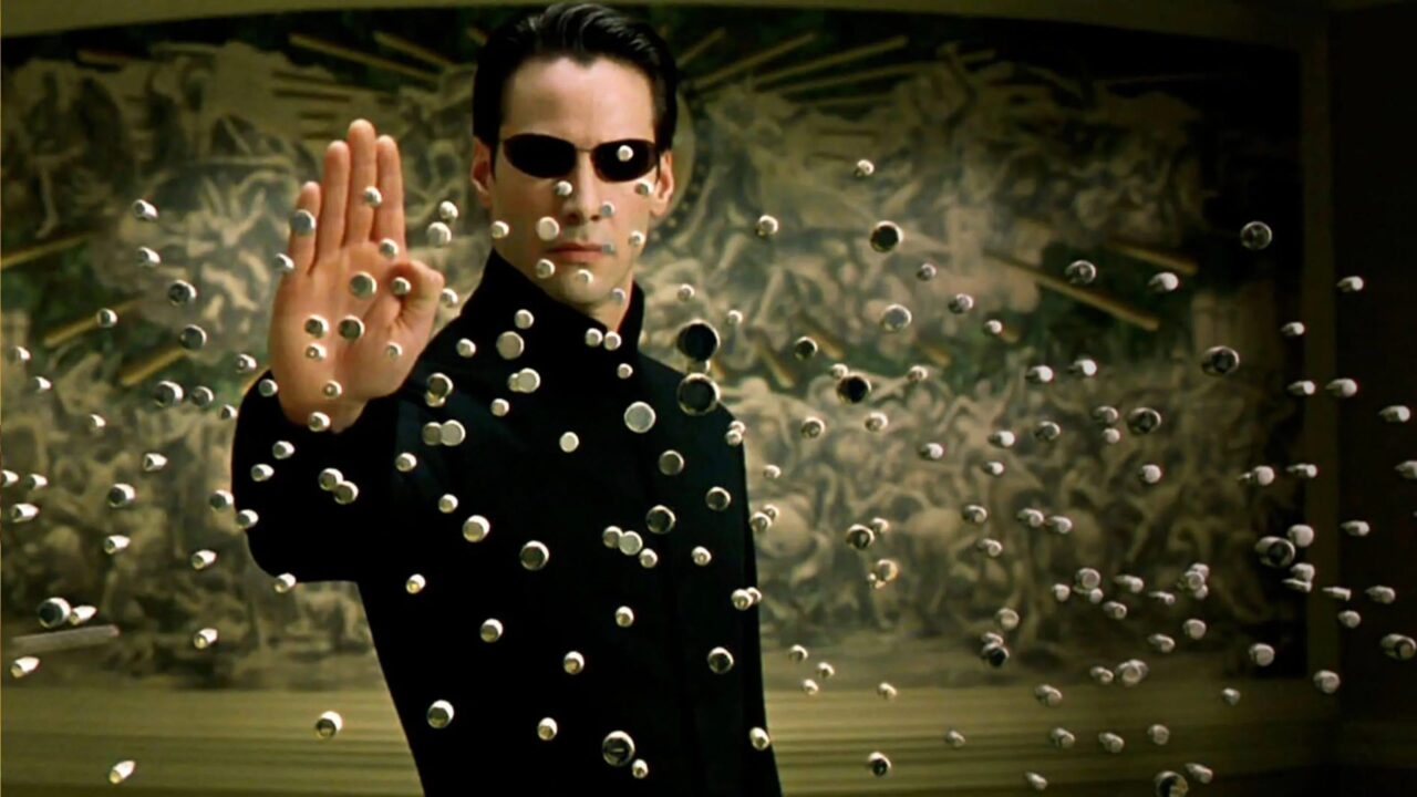Kadr z filmu Matrix. Mężczyzna w czarnym garniturze i okularach przeciwsłonecznych wyciąga rękę, zatrzymując w powietrzu kulki w ruchu na tle rozmazanego wzoru.