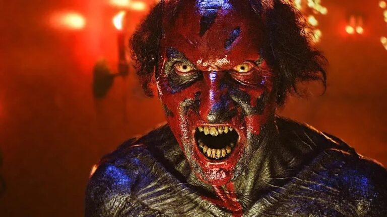 Osoba z teatralnym makijażem przedstawiająca demoniczny wygląd z rozwartymi ustami pokazującymi ostre zęby, mająca czerwoną skórę z czarnymi i niebieskimi akcentami oraz dzikie czarne oczy, na tle oświetlonym czerwoną poświatą.