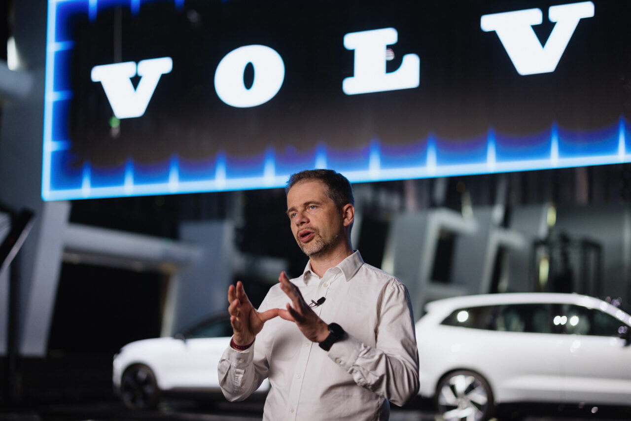 Mężczyzna w białej koszuli prezentujący i gestykulujący na scenie na tle niebieskiego oświetlenia z napisem "VOLVO" i białego samochodu.
