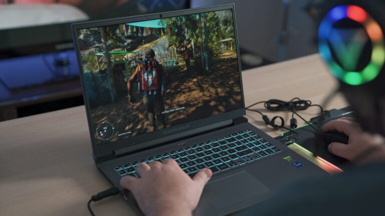 Laptop gamingowy z podświetlaną klawiaturą z włączoną grą. Urządzenie leży na biurku, ręka znajduje się w lewej części klawiatury