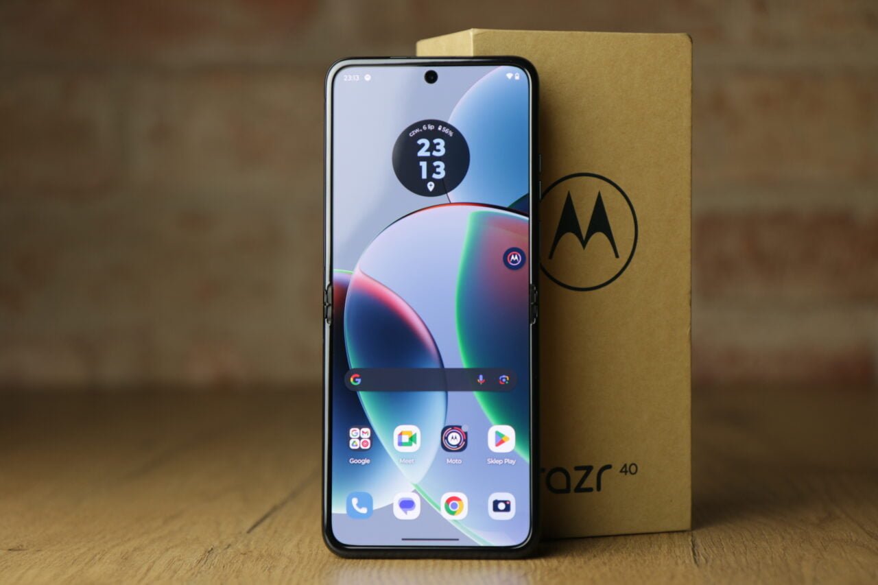 Smartfon marki Motorola stoi pionowo przed pudełkiem z logo producenta, na ekranie widoczny jest kolorowy interfejs użytkownika.