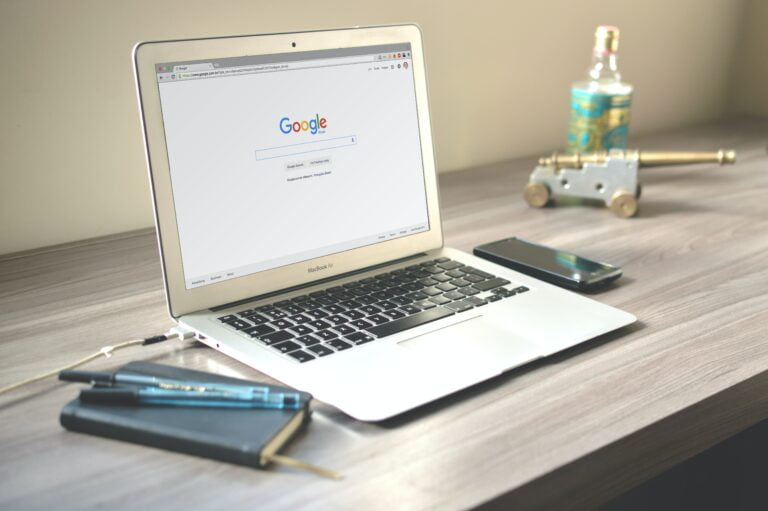 Laptop MacBook Air na drewnianym biurku z otwartą stroną główną wyszukiwarki Google, obok niego leżą długopisy, leży także smartfon, w tle niewyraźna butelka z płynem i mały model działa na kółkach.