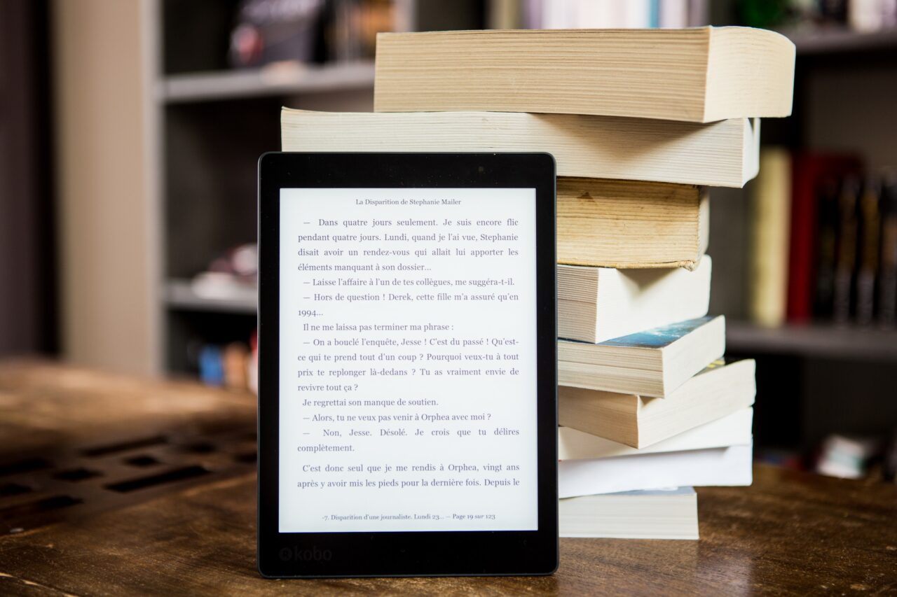Czytnik e-booków marki Kobo wyświetlający stronę z tekstem po francusku, oparty na stosie książek na drewnianym stole, z półkami z książkami w tle.