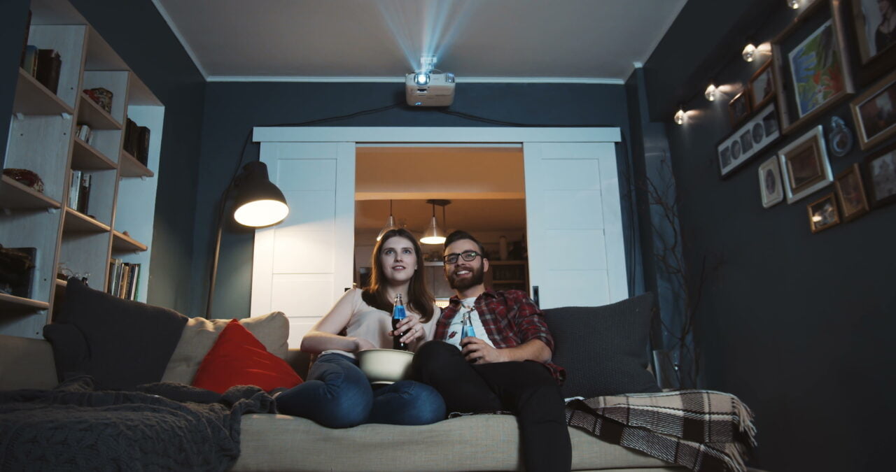 Mężczyzna i kobieta siedzący na kanapie w pokoju. Nad nimi wisi projektor wyświetlający obraz. Po obu stronach kadru regały z ksiażkami