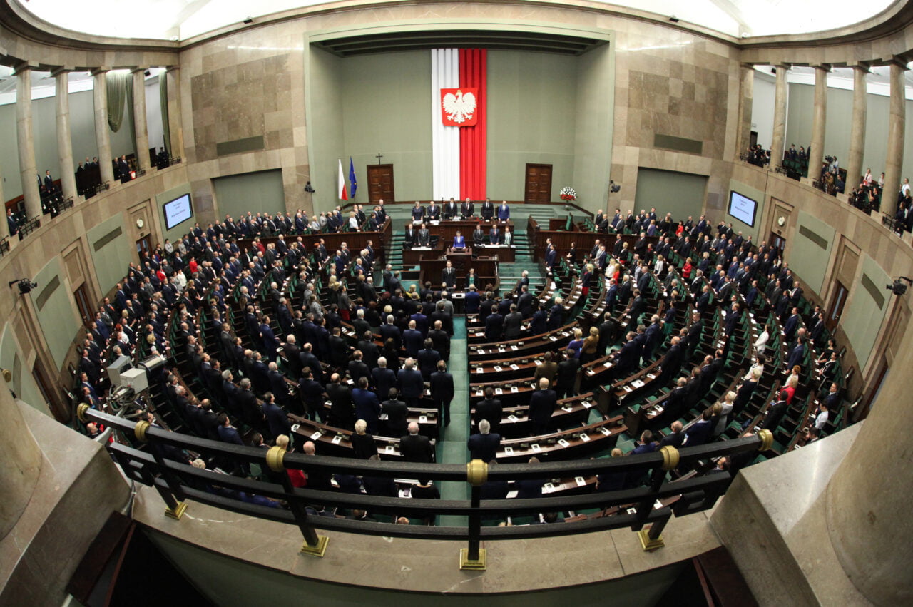 Widok wnętrza sali posiedzeń sejmu z posłami zgromadzonymi wokół mównicy, flaga Polski umieszczona na ścianie z tyłu.