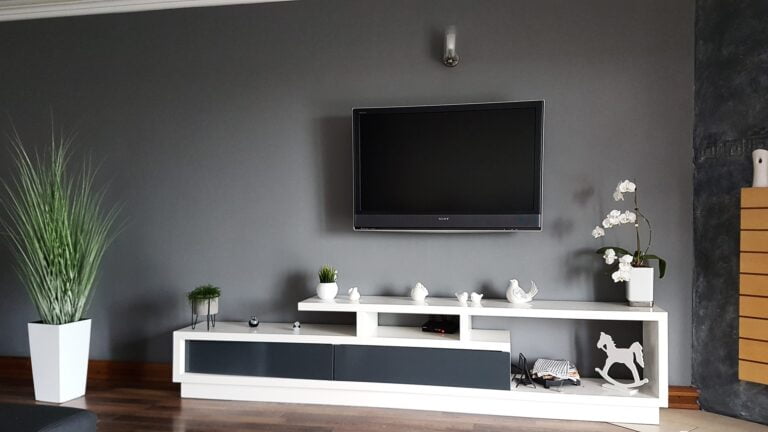 Nowoczesny salon z szarą ścianą, zawieszonym telewizorem, białą szafką RTV z dekoracjami i roślinami.