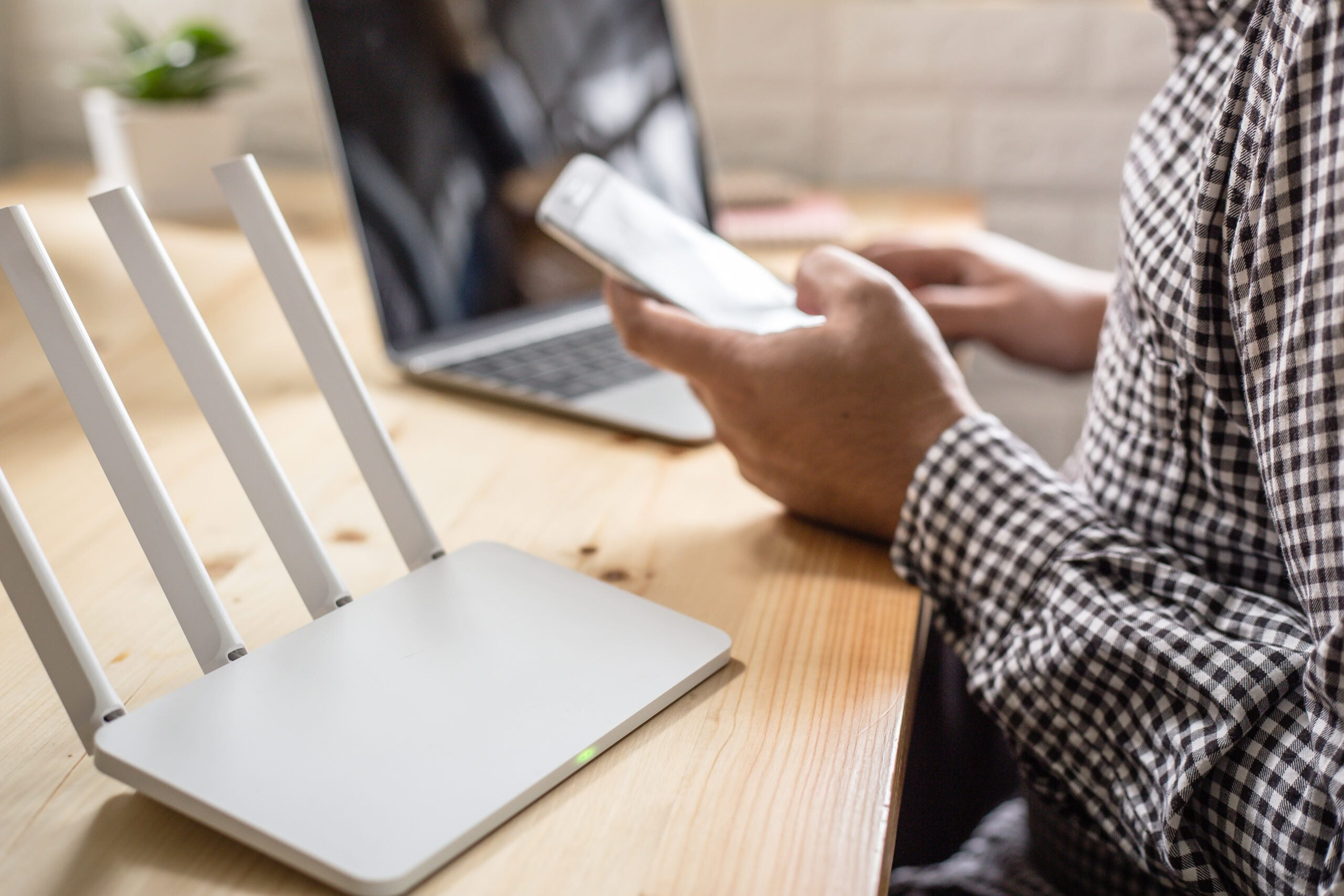 biały router z czterema antenami stojący na biurku, obok laptop, przed biurkiem mężczyzna siedzący z telefonem w ręce