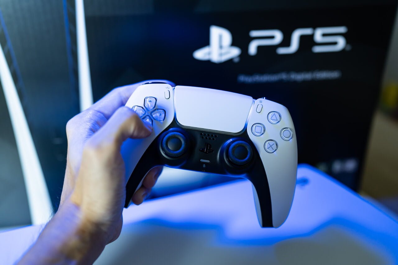 Ręka trzymająca biały kontroler do konsoli PlayStation 5 Pro z rozpoznawalnymi symbolami kierunków i przycisków na tle logo PS5.