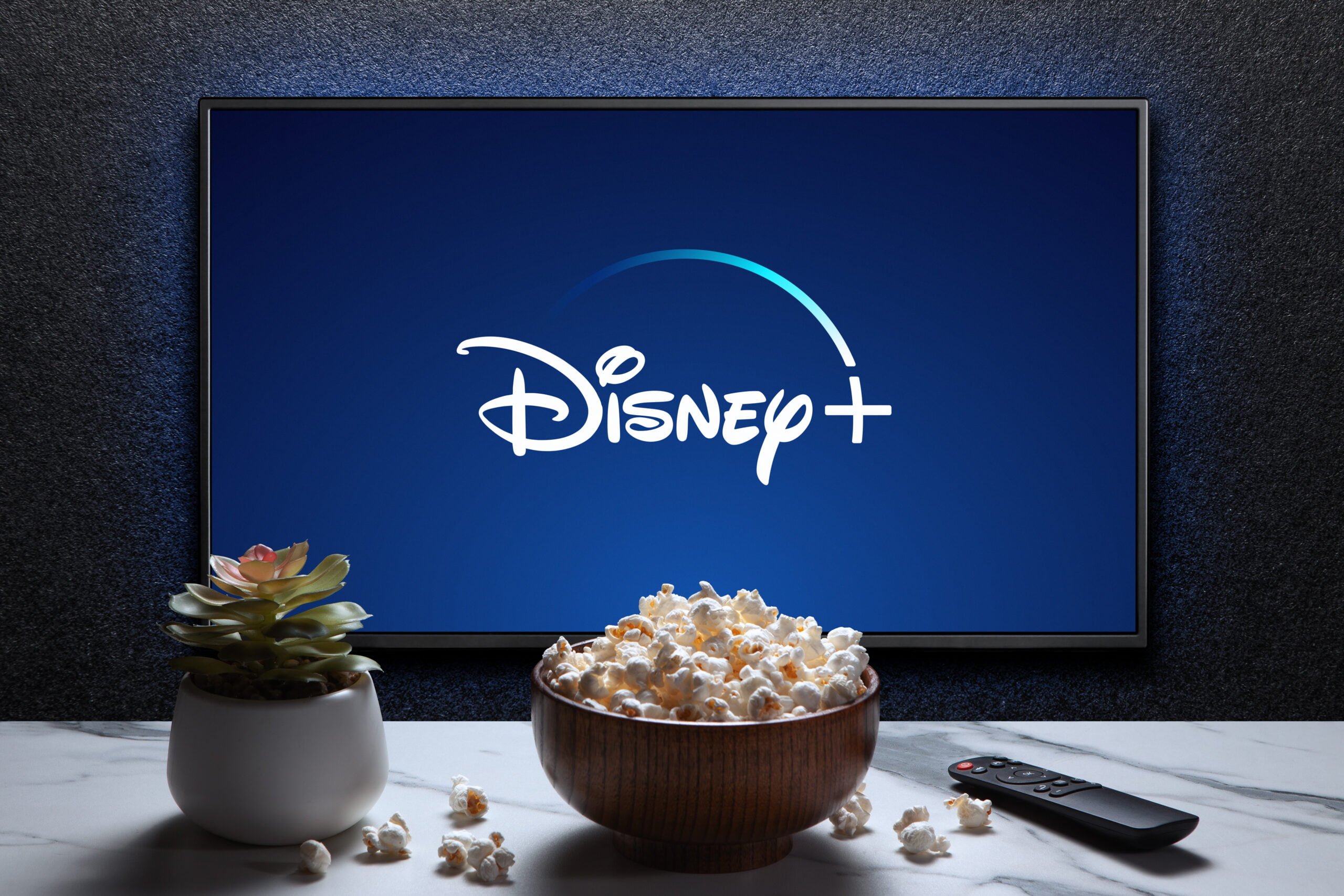 Telewizor wyświetlający logo Disney+, obok miseczka z popcornem, doniczka z sukulentem i pilot do telewizora na białym blacie.